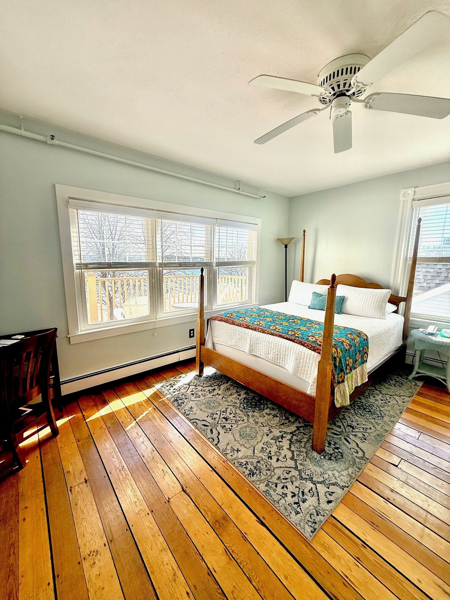 A bedroom with a four-poster bed, large windows, a ceiling fan, and hardwood flooring.