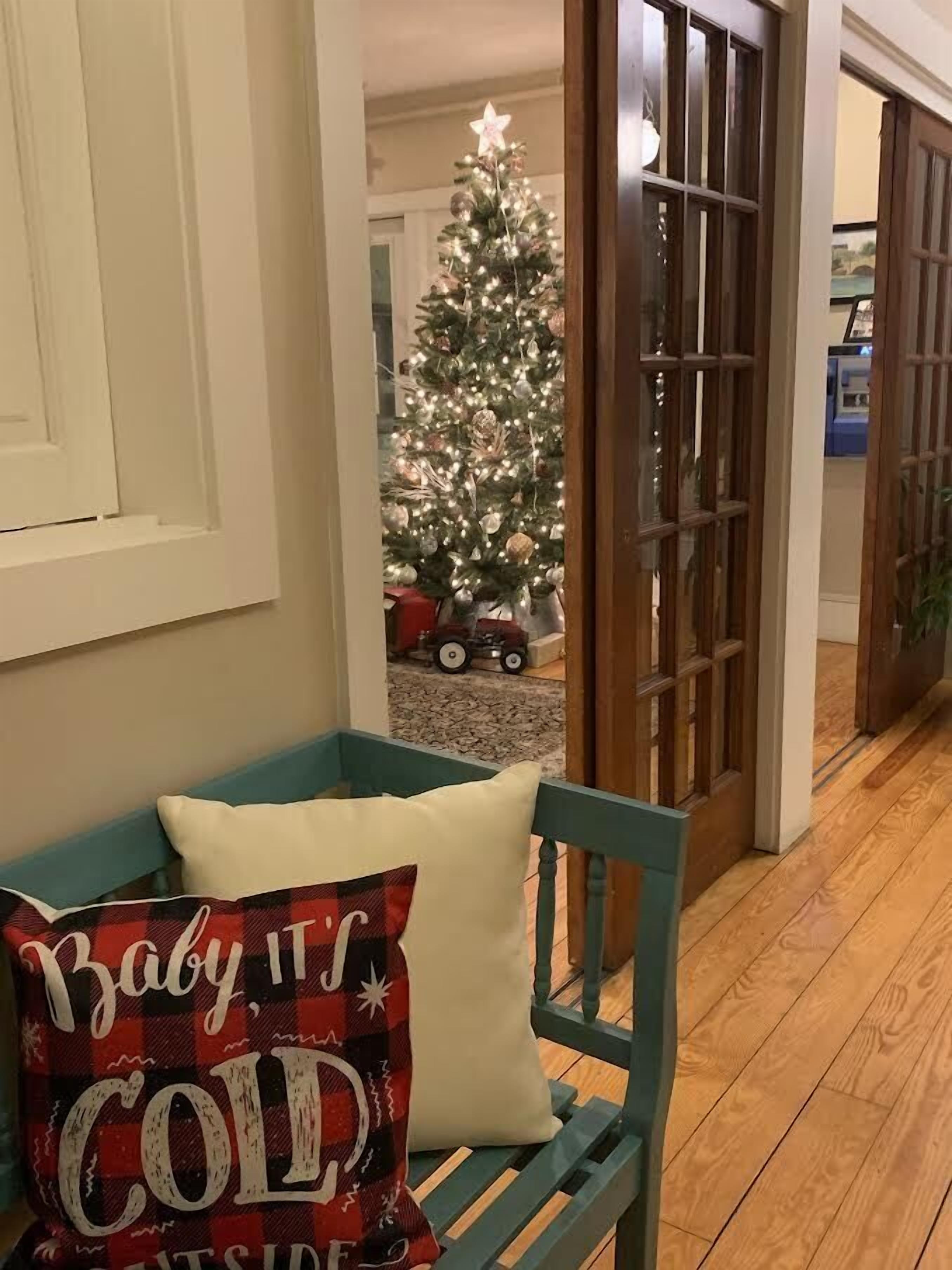 A teal bench with a white cushion and a pillow with "Baby it's cold" text. A Christmas tree with lights and ornaments is visible in the background.
