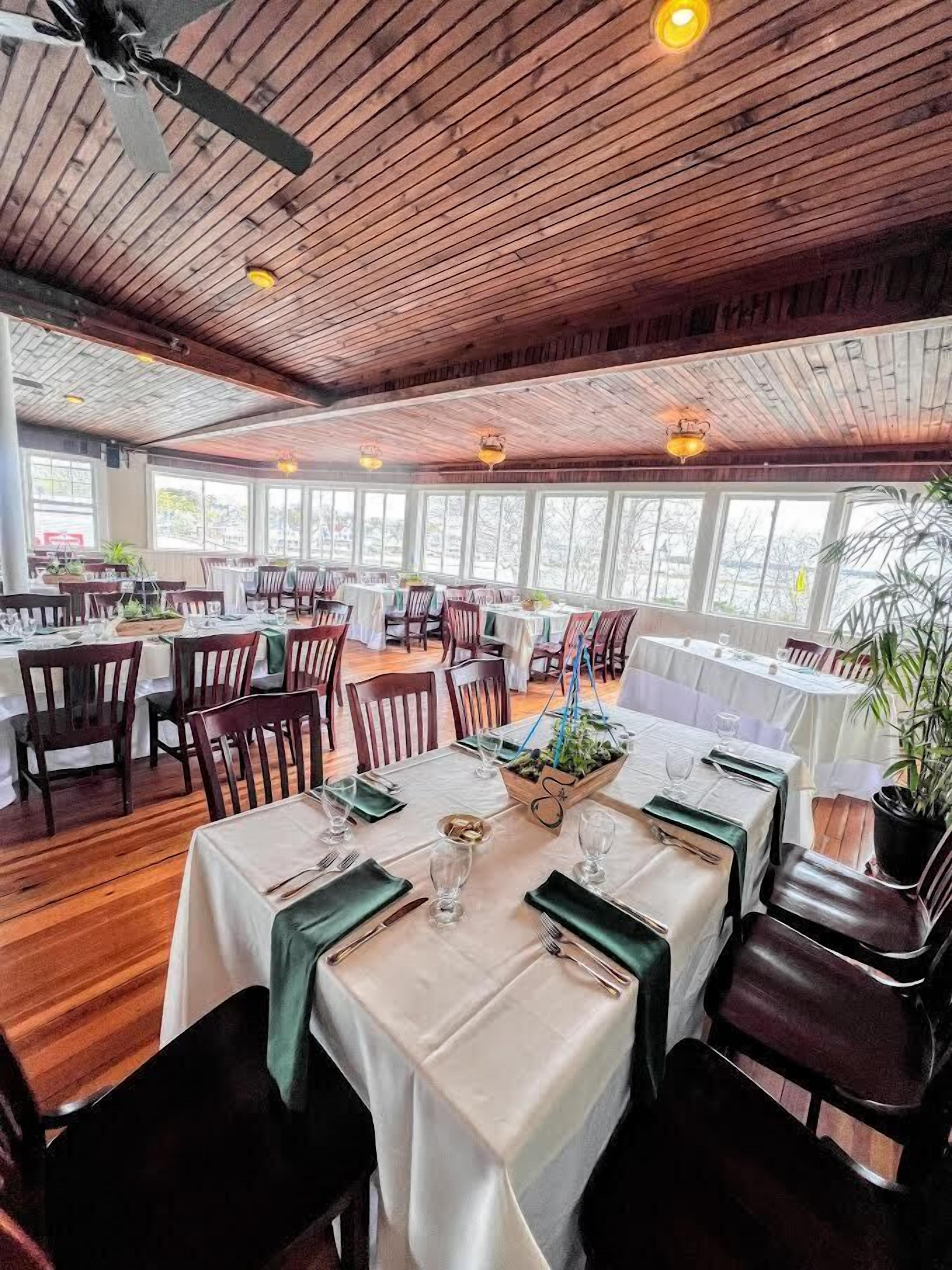 Restaurante com teto de madeira e janelas amplas. Mesas montadas com toalhas brancas e guardanapos verdes.