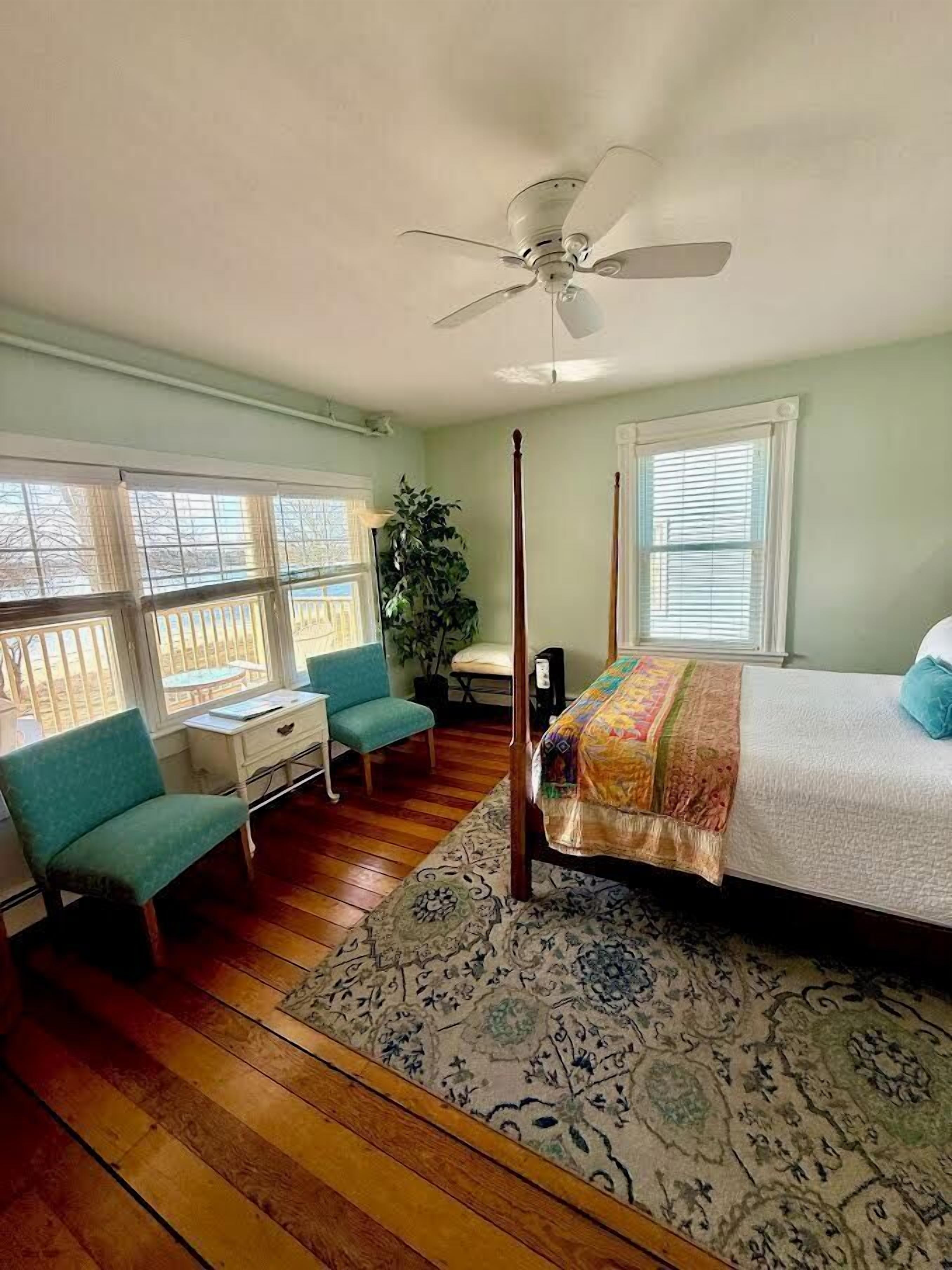 A bedroom with a four-poster bed, a ceiling fan, a window with blinds, a desk with a chair, and a large plant.