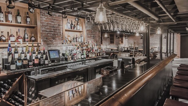 Un bar con una barra alargada, estanterías llenas de botellas y luces colgantes.