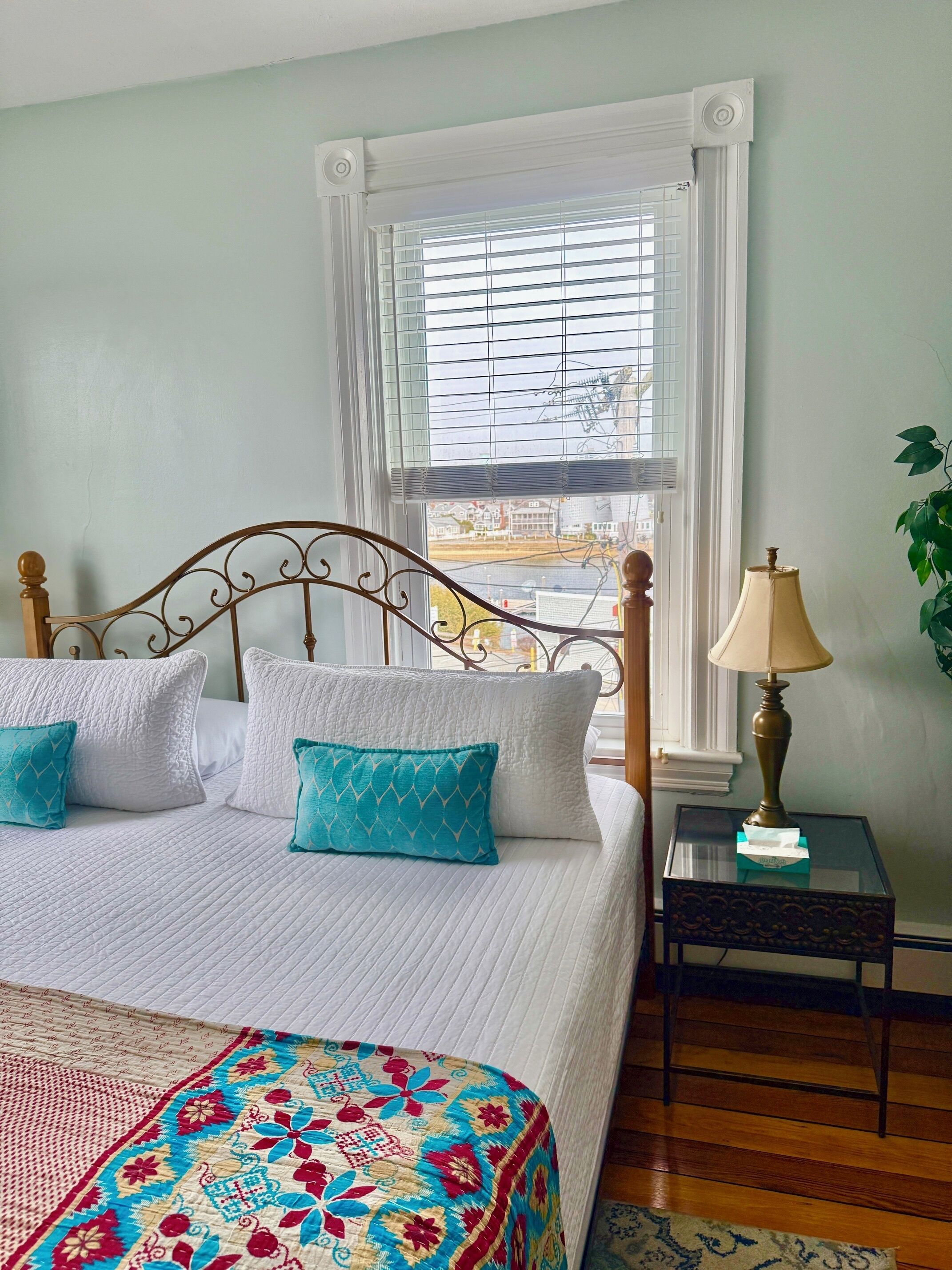 A neatly made bed with white and teal pillows, a patterned bedspread, a bedside lamp, and a view of the outside through a window with blinds.