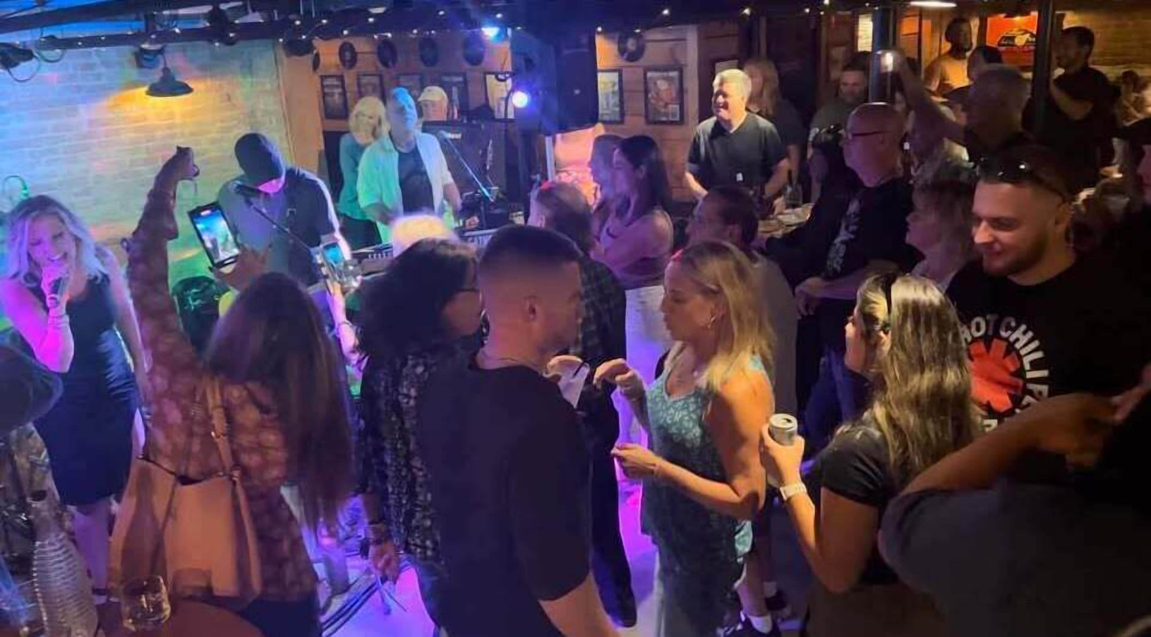 Un bar bondé où les gens discutent et s’amusent, avec une cabine de DJ en arrière-plan.