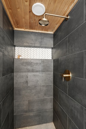 A shower with a wooden ceiling, a herringbone-patterned tile wall, and a gold showerhead.