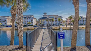 A wooden boardwalk leading to a building with a covered area, surrounded by palm trees and a body of water. - Carolina Dreamin - ocean, sandy beaches, restaurants, music and golfing (Myrtle Beach)