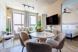Habitación de hotel moderna con sofá, dos sillones, una mesa redonda de comedor, un televisor de pantalla plana, cama y un ventanal grande con cortinas.