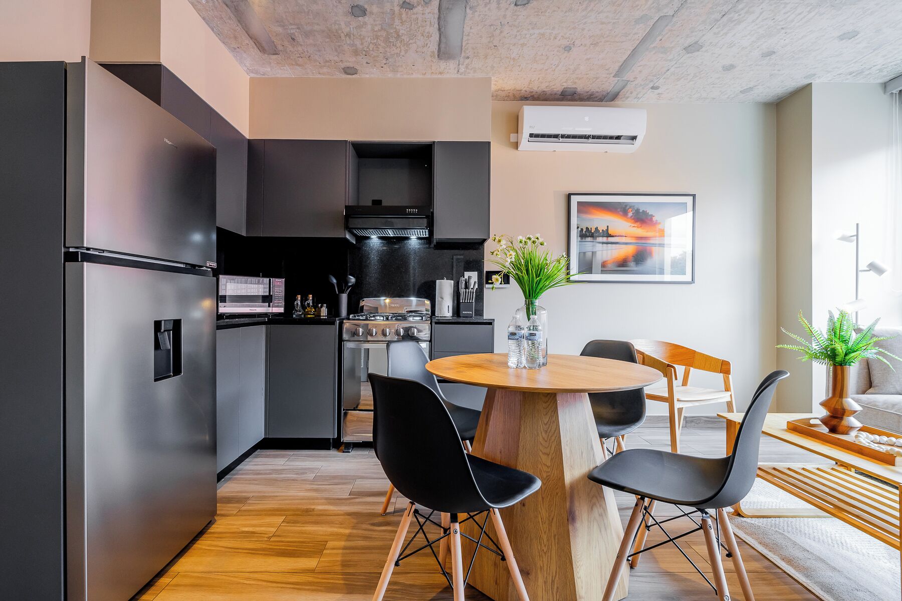 Eine moderne Küche mit Einbauofen und Herd, ein Essbereich mit einem runden Holztisch und schwarzen Stühlen sowie ein Wohnbereich mit einer Couch und einer Pflanze.