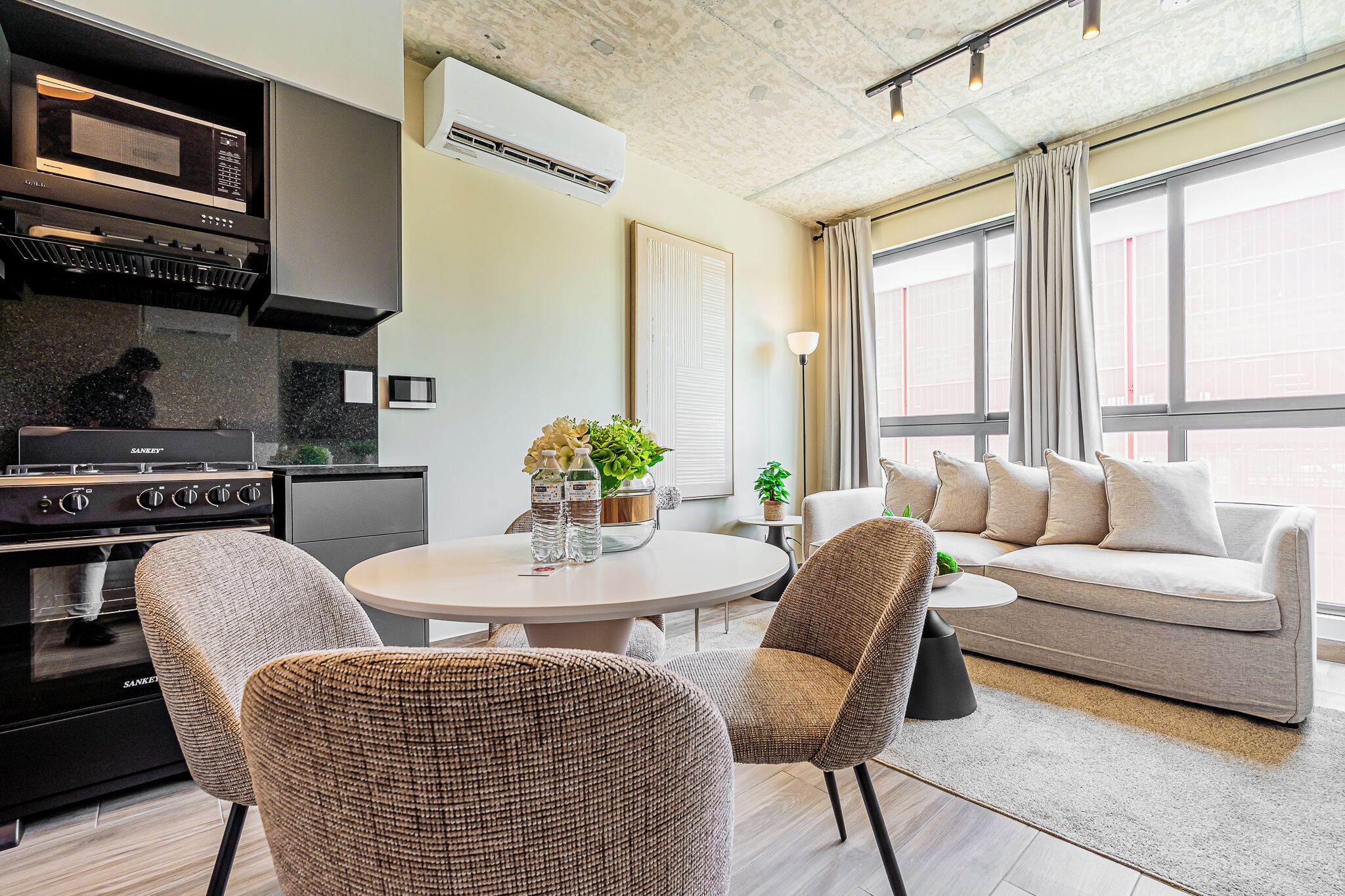 Une cuisine moderne équipée d’un four à micro-ondes et d’une cuisinière intégrés, un coin repas avec une table ronde et des chaises, et un salon avec un canapé et une vue sur la ville grâce à de grandes fenêtres.