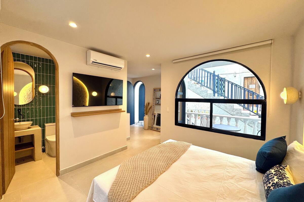 A modern hotel room with a large bed, a bathroom with a green-tiled wall, a large window with a view of a staircase, and a mounted artwork above the bed.