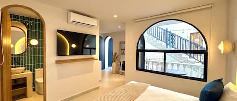 A modern hotel room with a large bed, a bathroom with a green-tiled wall, a large window with a view of a staircase, and a mounted artwork above the bed.