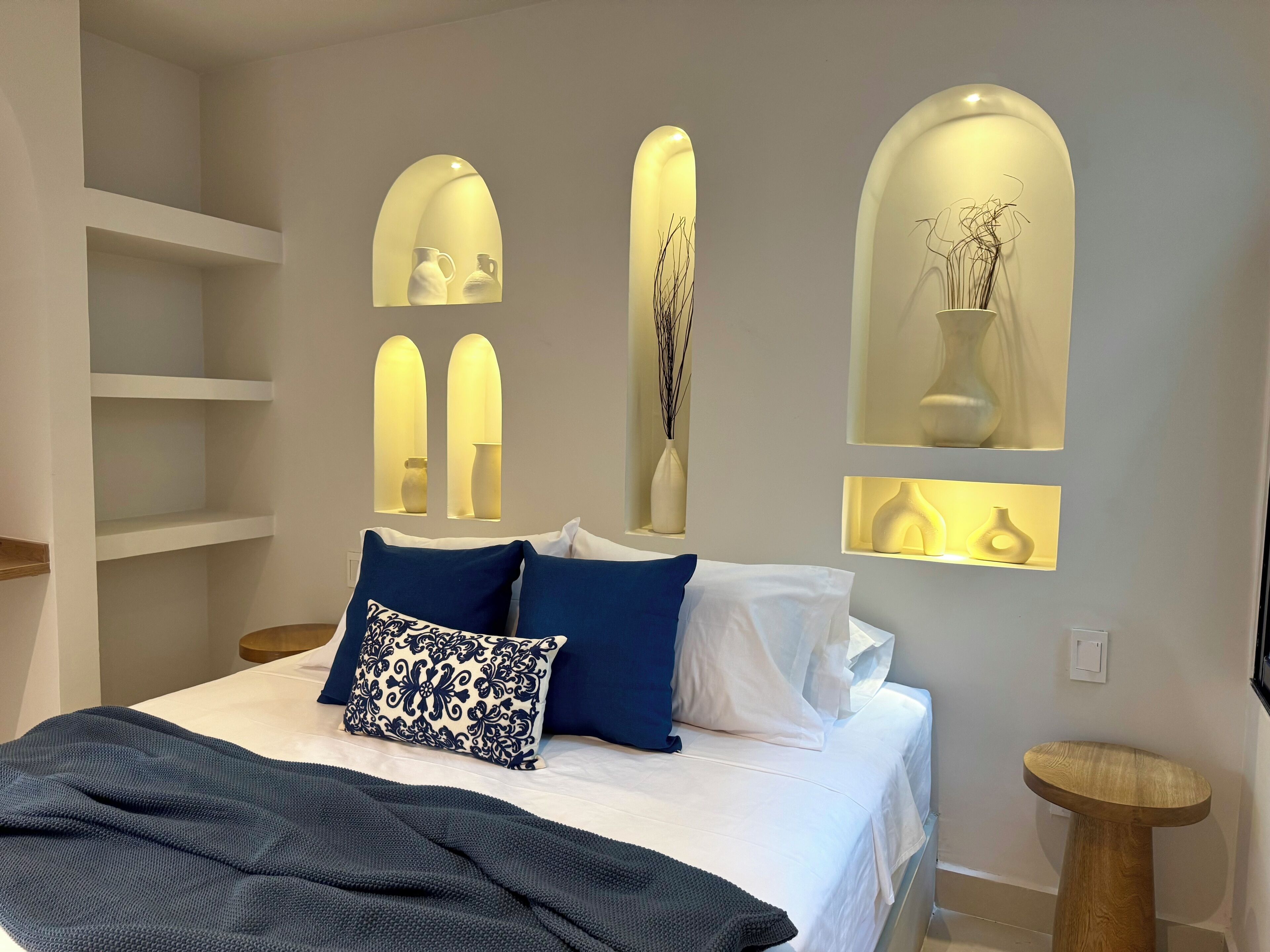 A bedroom with a bed, pillows, a bedside table, and wall-mounted shelves with vases.