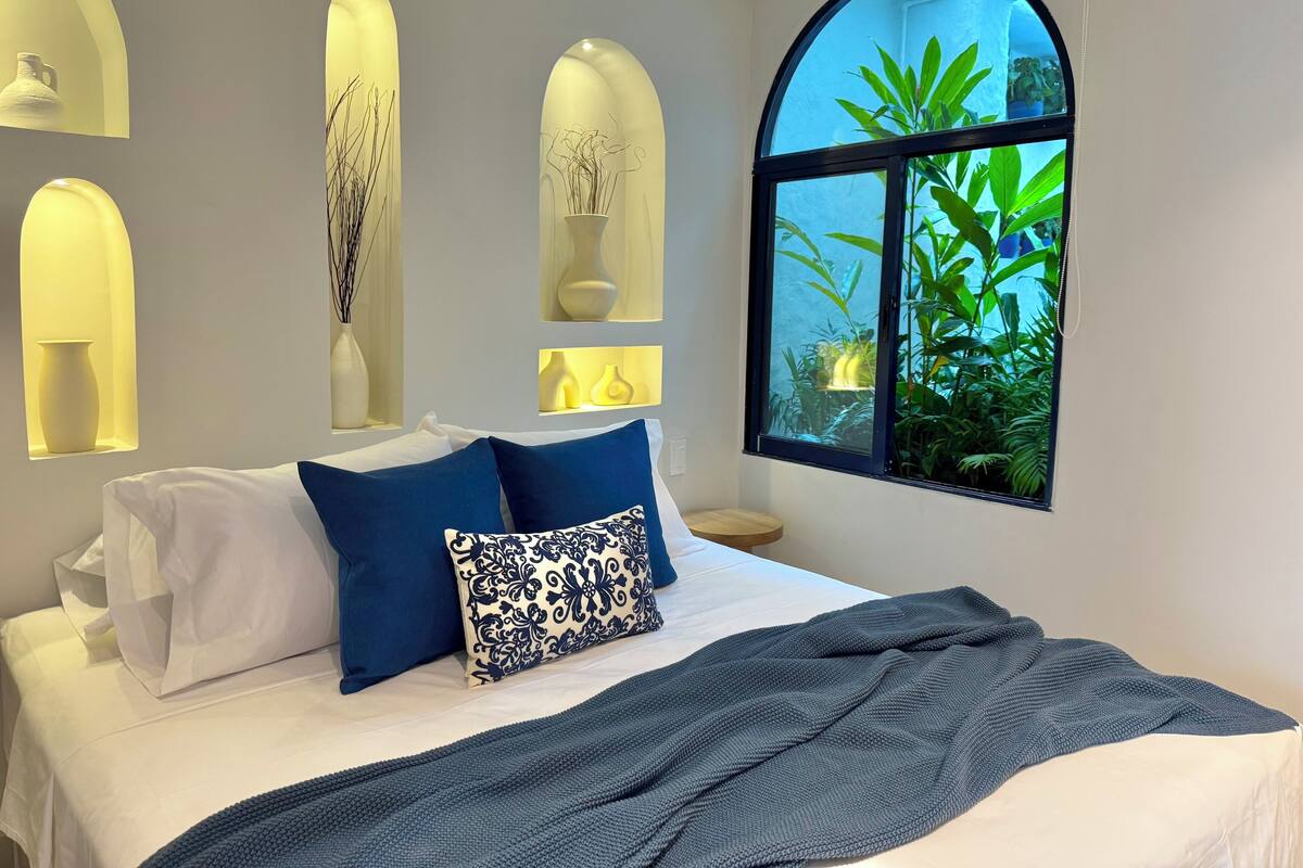 A bedroom with a bed, pillows, a bedside table, and a window with a view of greenery.