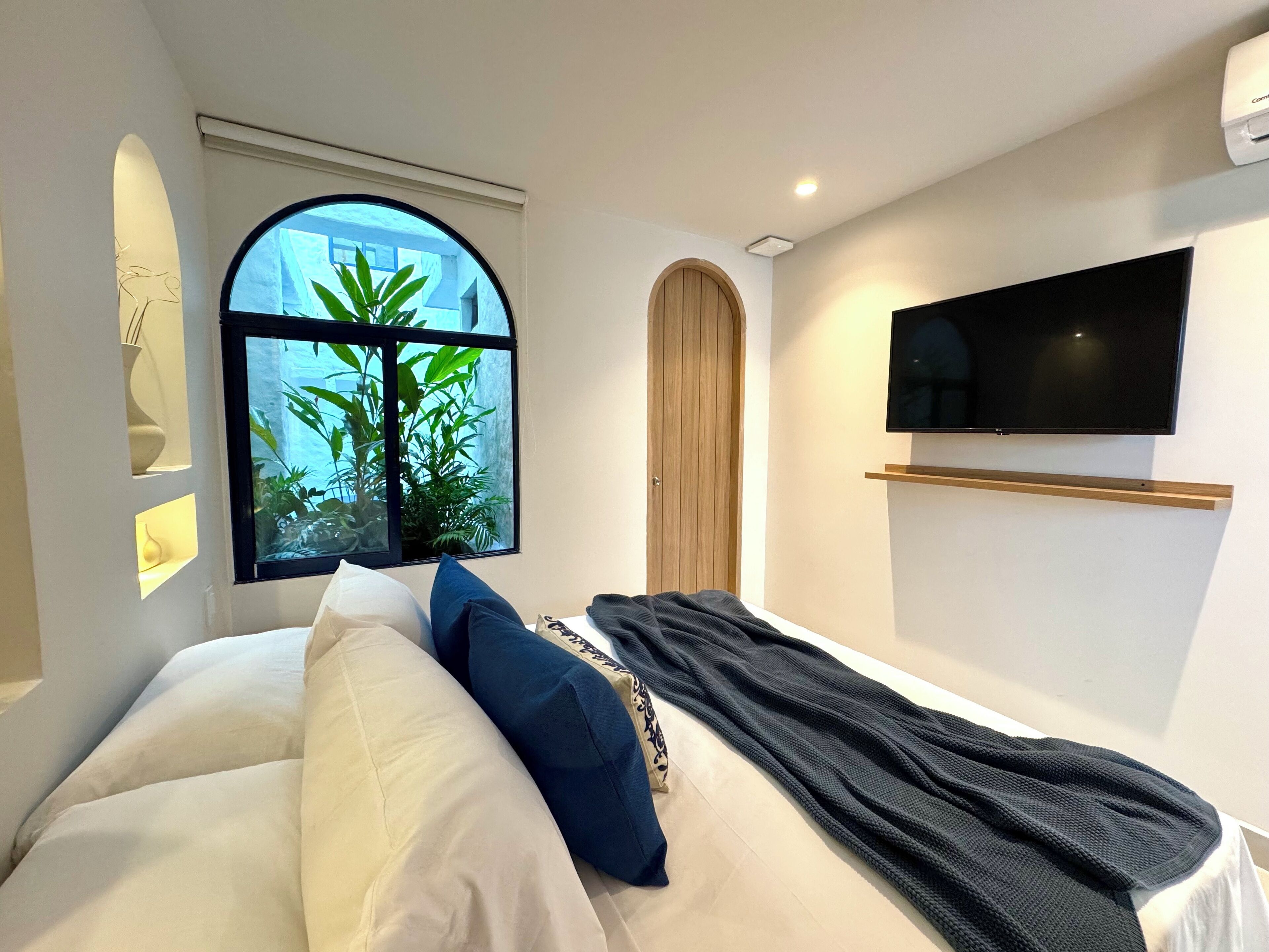 A modern bedroom with a large bed, a TV mounted on the wall, and a window with a view of greenery.