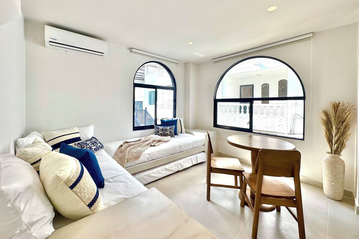 A modern bedroom with a bed, two windows, a round table, and a chair.