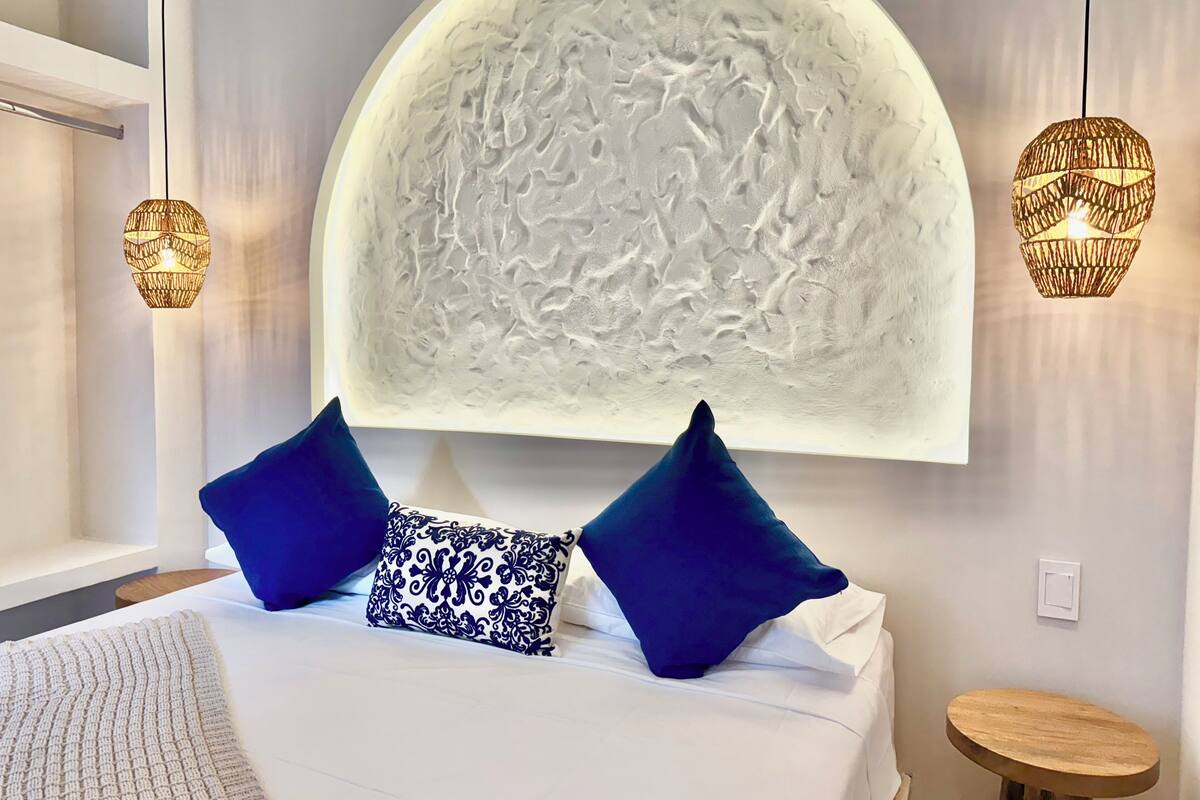 A bedroom with a large, textured wall art piece above the bed, two hanging lamps, and a small wooden table beside the bed.