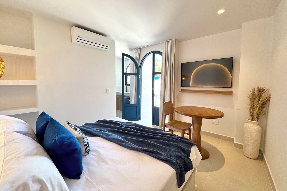 A modern hotel room with a bed, a round wooden table, a chair, a TV mounted on the wall, and a large window with curtains.