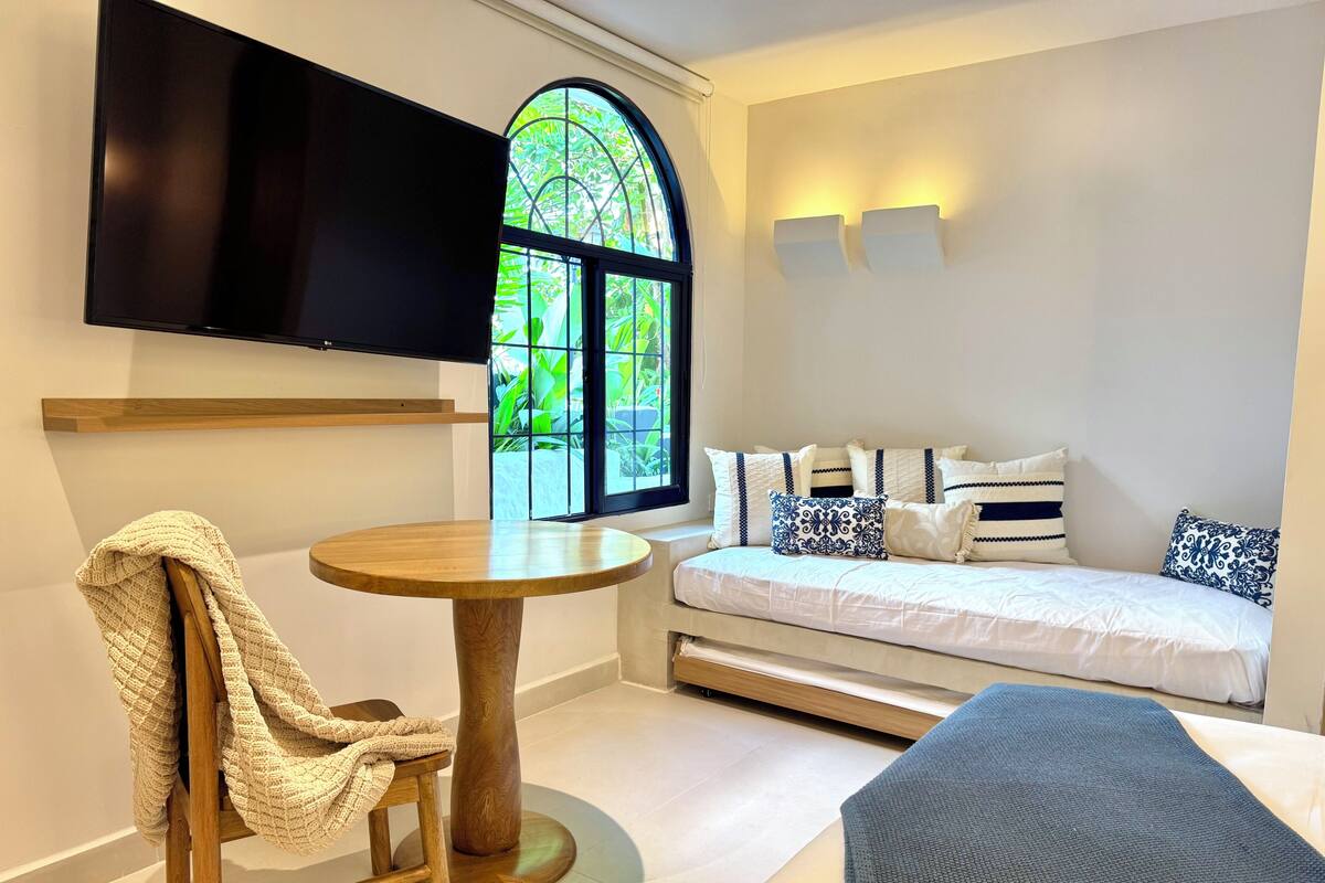 A modern room with a round wooden table, a sofa with patterned cushions, a TV mounted on the wall, and a large arched window with greenery outside.