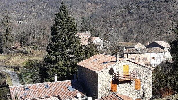 A stone building with a tiled roof, a wooden balcony, and a view of a valley and mountains in the background.