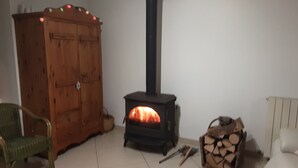 A room with a wooden cabinet, a black stove with a chimney, a chair, and a stack of firewood.