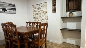 A dining area with a wooden table and chairs, a stone wall, a hanging light fixture, and a wooden shelf with a basket. - La Cuadra. Peace and tranquility in a privileged rural setting (Villaluenga del Rosario)