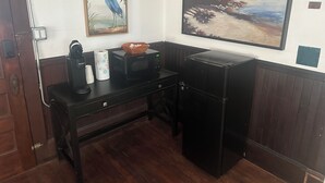 A compact room with a wooden desk, a black refrigerator, a painting of a bird, and a painting of a landscape.