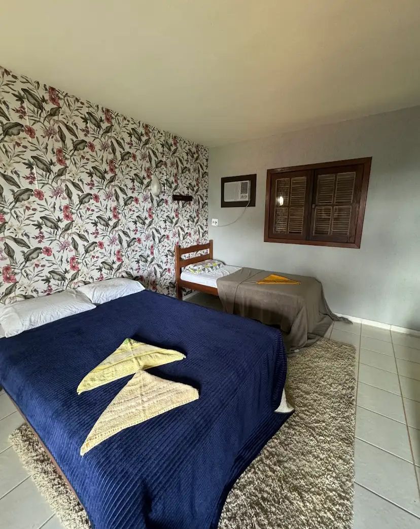 Un dormitorio con una cama grande, una cama más pequeña, papel tapiz floral, un equipo de aire acondicionado y una ventana con contraventanas.
