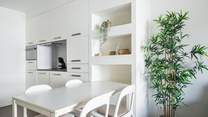 A modern kitchen with white cabinetry, a built-in oven, and a dining area with a white table and chairs. There is a large potted bamboo plant on the right side. - GuestReady - Welcoming place in Porto (Boavista)