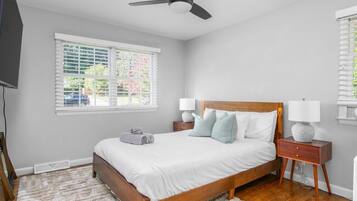 A bedroom with a bed, bedside lamps, a ceiling fan, a window with blinds, and a view of greenery outside.