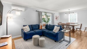 Ein modernes Wohnzimmer mit einer Eckcouch, einem Esstisch mit Stühlen und einem großen Fenster mit Vorhängen.