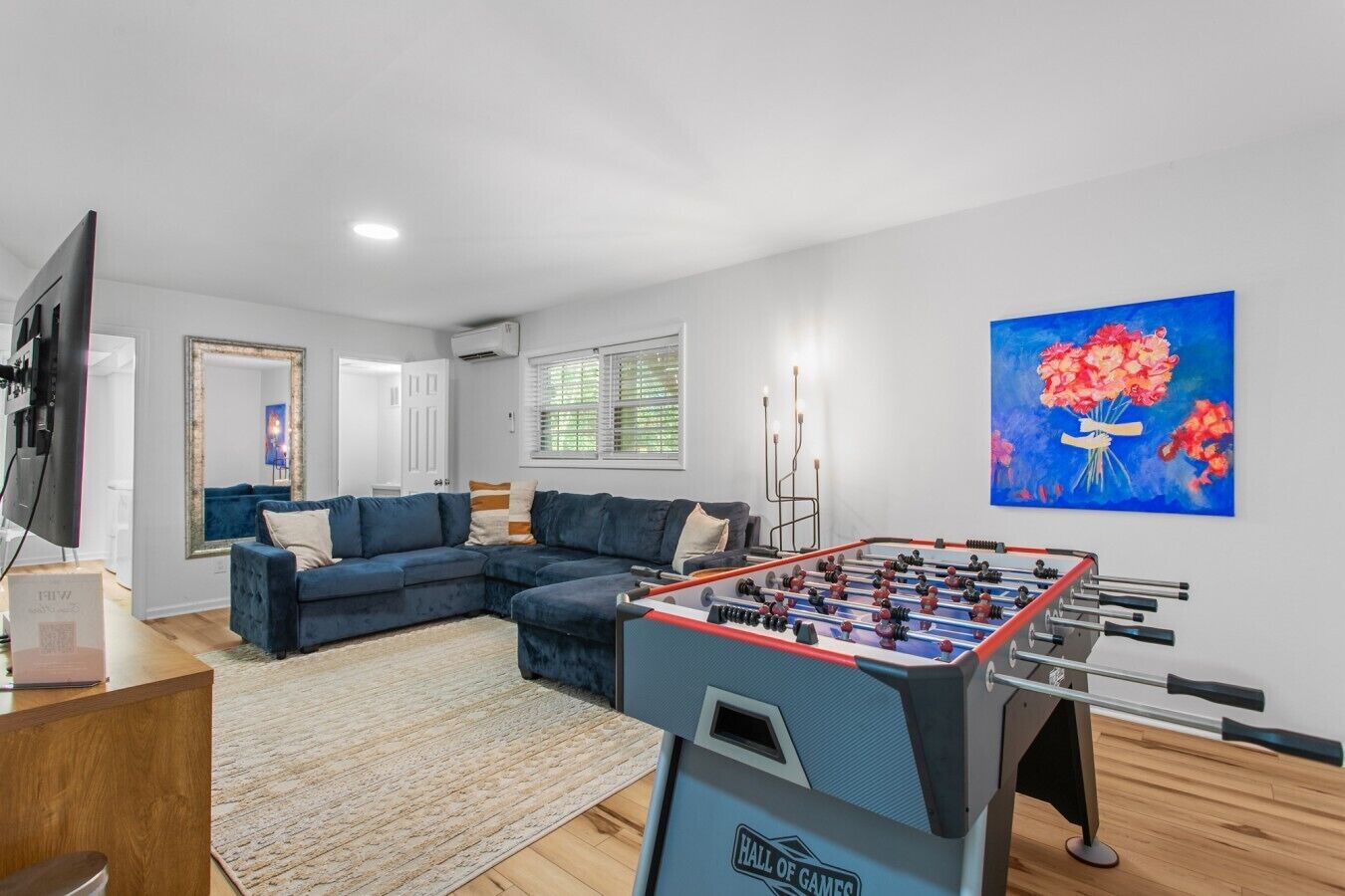 A living room with a foosball table, a blue sofa, a television, and a painting of flowers.