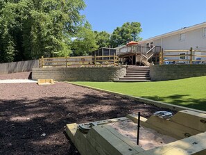 Un patio trasero con terraza de madera, muro de piedra y zona cubierta de césped.