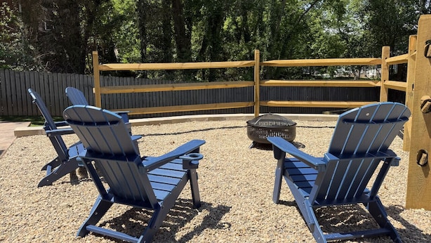 Zwei Adirondack-Stühle in einem Kiesbereich mit Holzlattenzaun und Bäumen im Hintergrund.