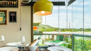 A dining area with a table set for two, a bottle of champagne, and a view of a green landscape through large windows. - Flat 208-C  Condomínio Carneiros Resort (Tamandaré)