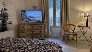 A bedroom with a bed, a dresser, a television displaying a beach scene, a chair, and a small table with a lamp. - Le 52 en Provence (Robion)