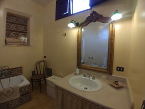 Une salle de bain avec un lavabo blanc, un miroir doté d’un cadre doré orné et une fenêtre située au-dessus du lavabo.