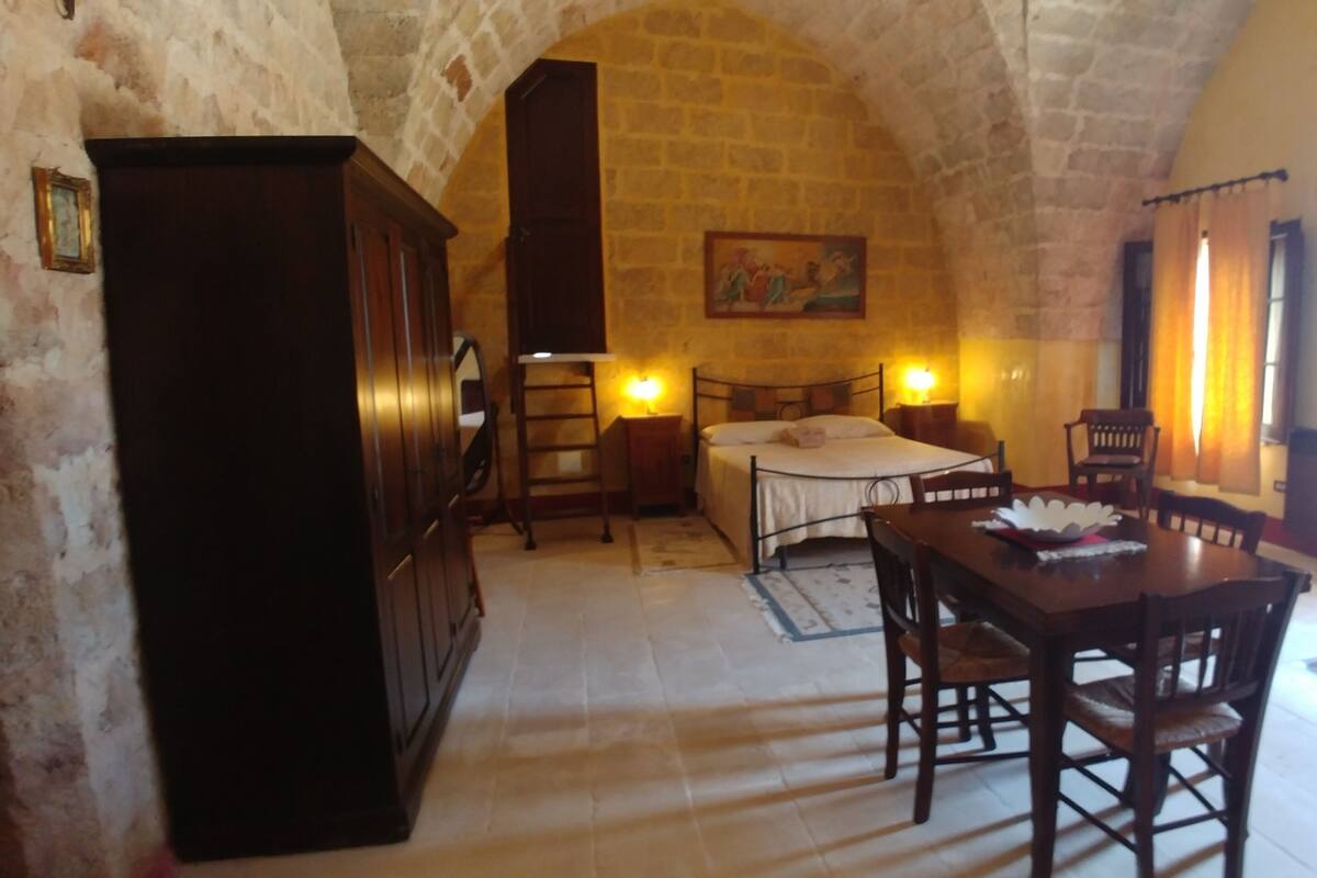 A room with a stone arch ceiling, a bed with a white bedspread, a wooden table with chairs, and a painting on the wall.