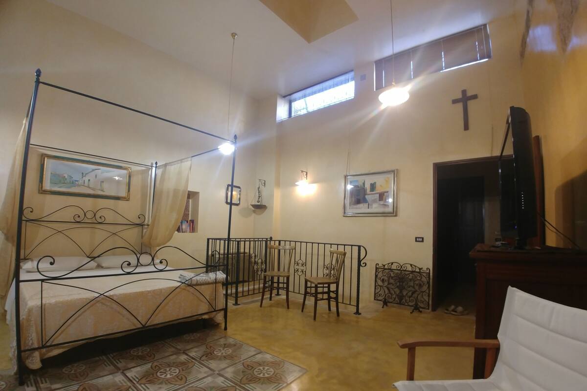 A bedroom with a four-poster bed, a television, a chair, and a window with a cross on the wall.