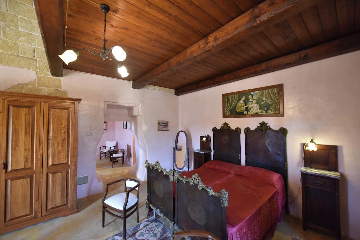 A bedroom with a wooden ceiling, a bed with a red bedspread, a wooden wardrobe, a chair, and a small table.