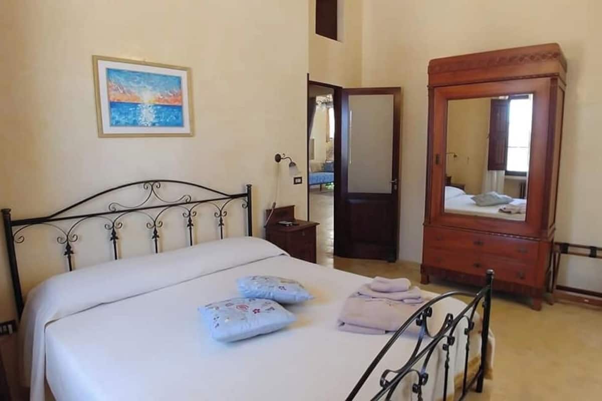 A bedroom with a bed, bedside table, dresser, mirror, and a door leading to another room.