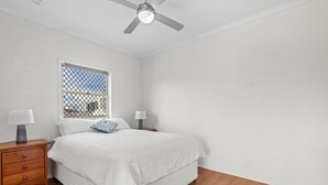 Une chambre à coucher avec un lit, deux tables de chevet avec des lampes, un ventilateur de plafond et une fenêtre munie d’un moustiquaire métallique.