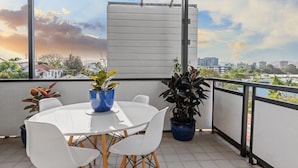 Un balcon avec une table ronde et des chaises, deux plantes en pot et une vue sur le paysage urbain.