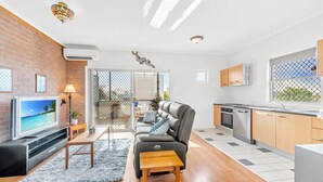 Un appartement moderne doté d’un salon comprenant un canapé, une télévision et une petite table en bois. La cuisine est équipée de meubles en bois et d’un lave-vaisselle.