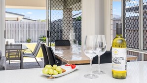 Et bord med en vinflaske, to glas, en tallerken med druer og vandmelon, og udsigt til en baghave gennem et insektnetdør.