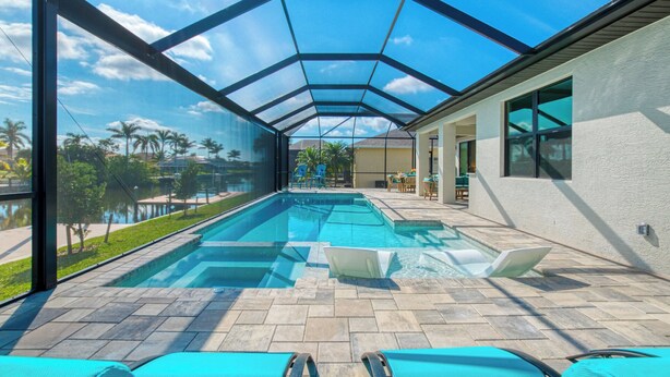 A swimming pool with a glass ceiling, lounge chairs, and a view of palm trees and a lake. - VRCC Villa Tiki Oasis (Cape Coral)