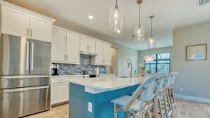 A modern kitchen with stainless steel appliances, a blue island, wicker bar stools, and pendant lighting. - VRCC Villa Tiki Oasis (Cape Coral)