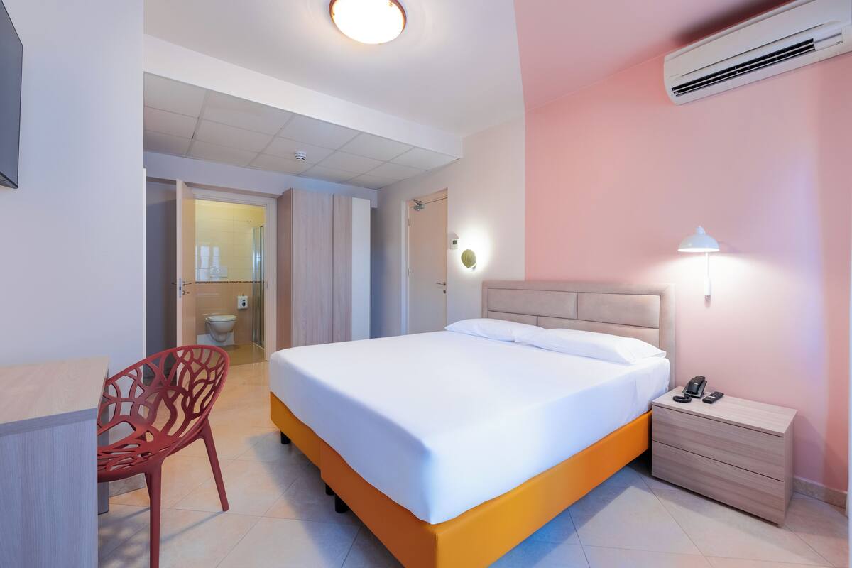 A modern hotel room with a large bed, a desk, a chair, a bedside table with a lamp, and an air conditioning unit.