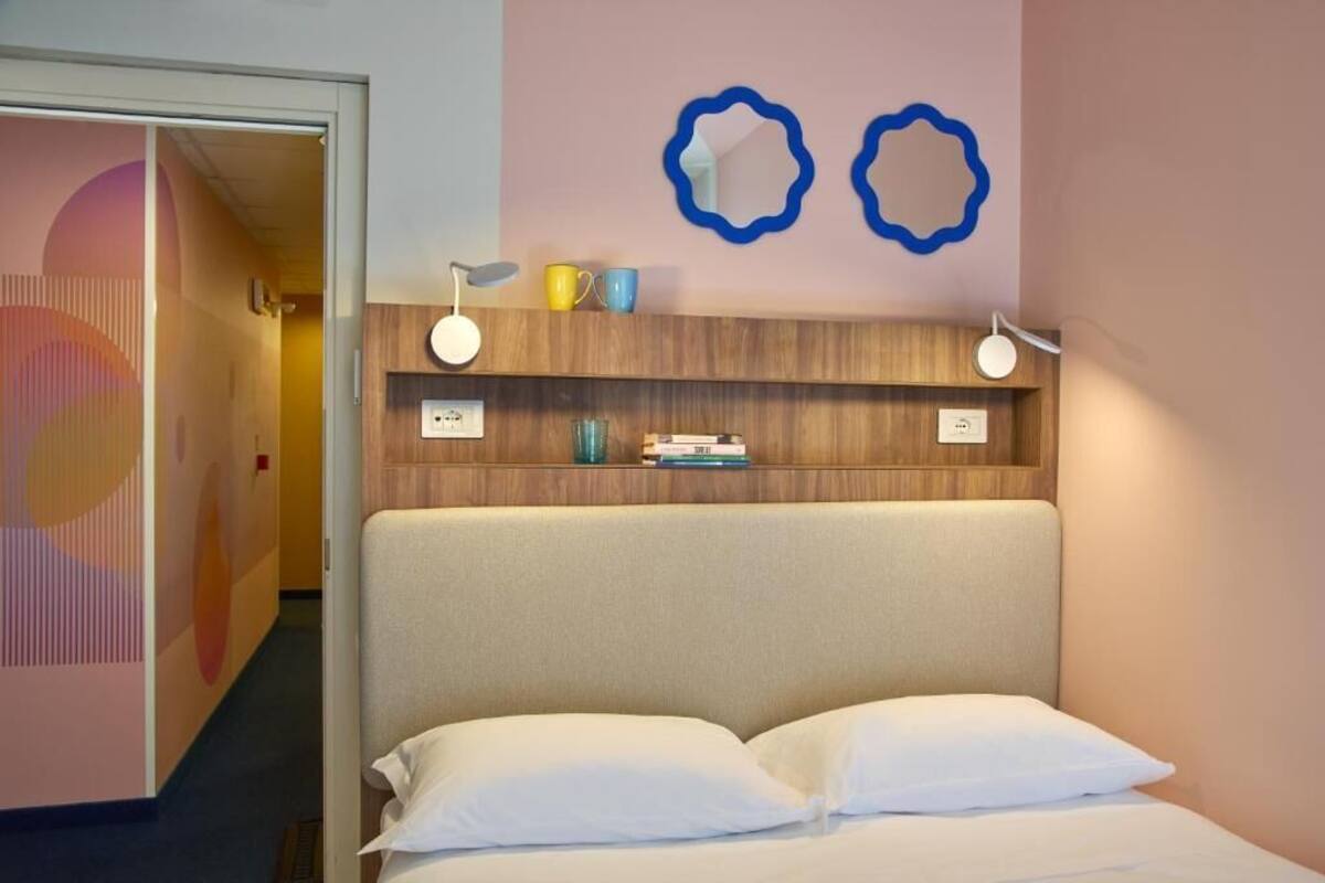 A hotel room with a bed, a wooden headboard, a shelf with books and cups, and two wall mirrors.