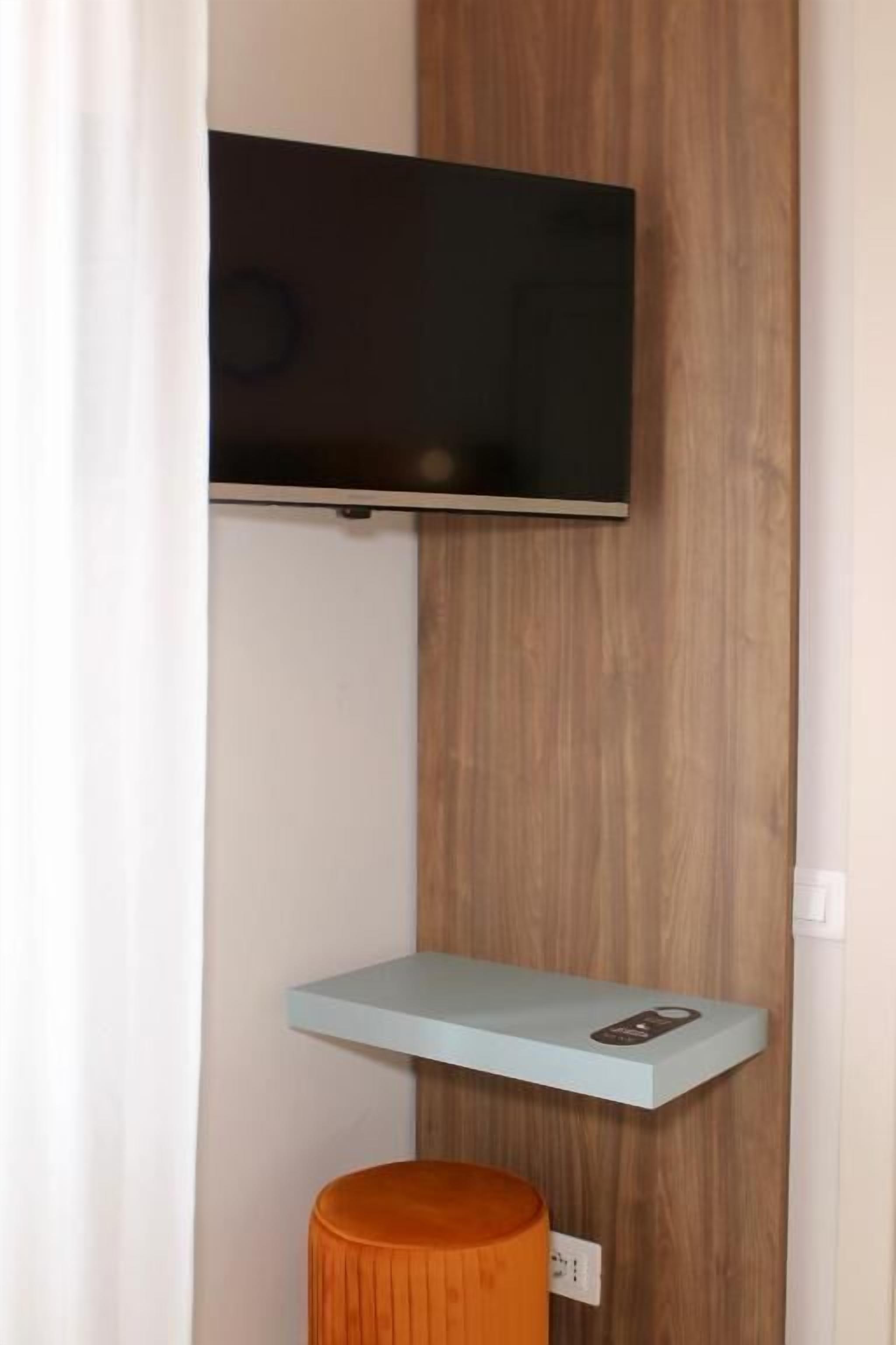 A room with a mounted TV on a wooden wall, a light blue shelf below it, and an orange stool.