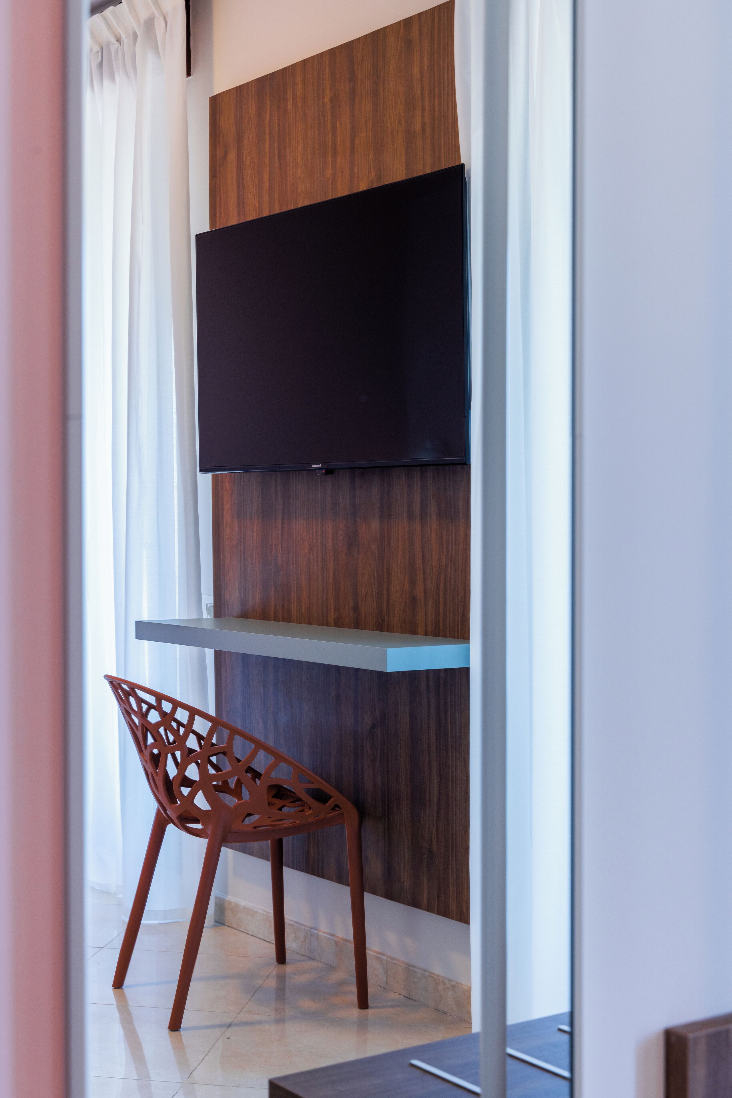 A modern interior with a flat-screen TV mounted on a wooden panel wall, a metal shelf below, and a unique red chair with a geometric design.