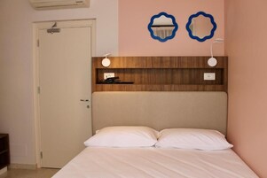 Un dormitorio con cama, una puerta, una estantería de madera con objetos y dos luces fijadas a la pared.